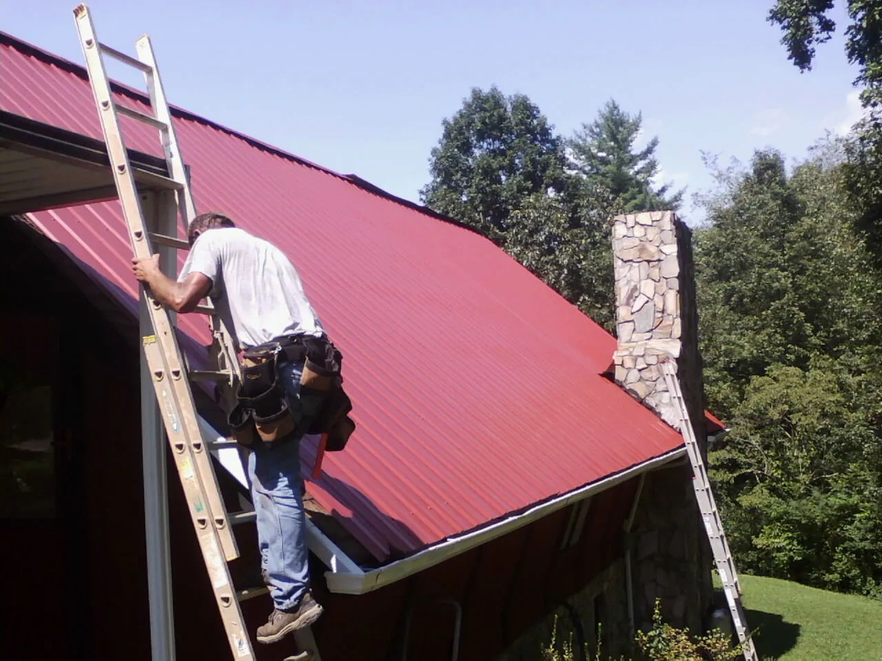 Roofer on a ladder beside a red metal roof and stone chimney — Soffit & Fascia Repair by Roofing Brothers in River Rouge